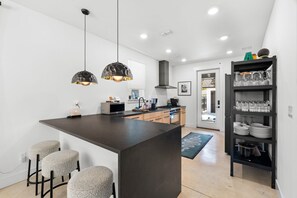A modern kitchen with a central island, bar stools, pendant lighting, and a glassware shelf. - The Slate Retreat | Pool + Hot Tub (Jacksonville)