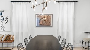 Een moderne eetruimte met een donkere tafel, zwarte draadstoelen, een plafondlamp en een lijstwerk met abstracte kunst.