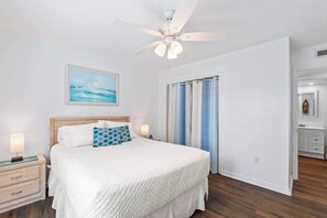 A bedroom with a bed, a ceiling fan, a nightstand, a bedside lamp, a painting, and a bathroom door. - Salt + Sun | Beach + Pool (St. Augustine)