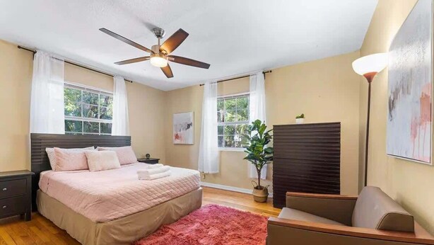 A bedroom with a bed, a ceiling fan, a window, a plant, and a nightstand. - Eclectic Escape #1 | Team Joseph Ellen (Jacksonville)