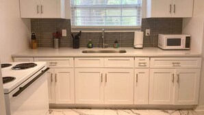 A kitchen with white cabinets, a stove, a microwave, and a window with blinds. - Eclectic Escape #1 | Team Joseph Ellen (Jacksonville)