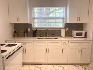 A kitchen with white cabinets, a stove, a microwave, and a window with blinds. - Eclectic Escape #1 | Team Joseph Ellen (Jacksonville)