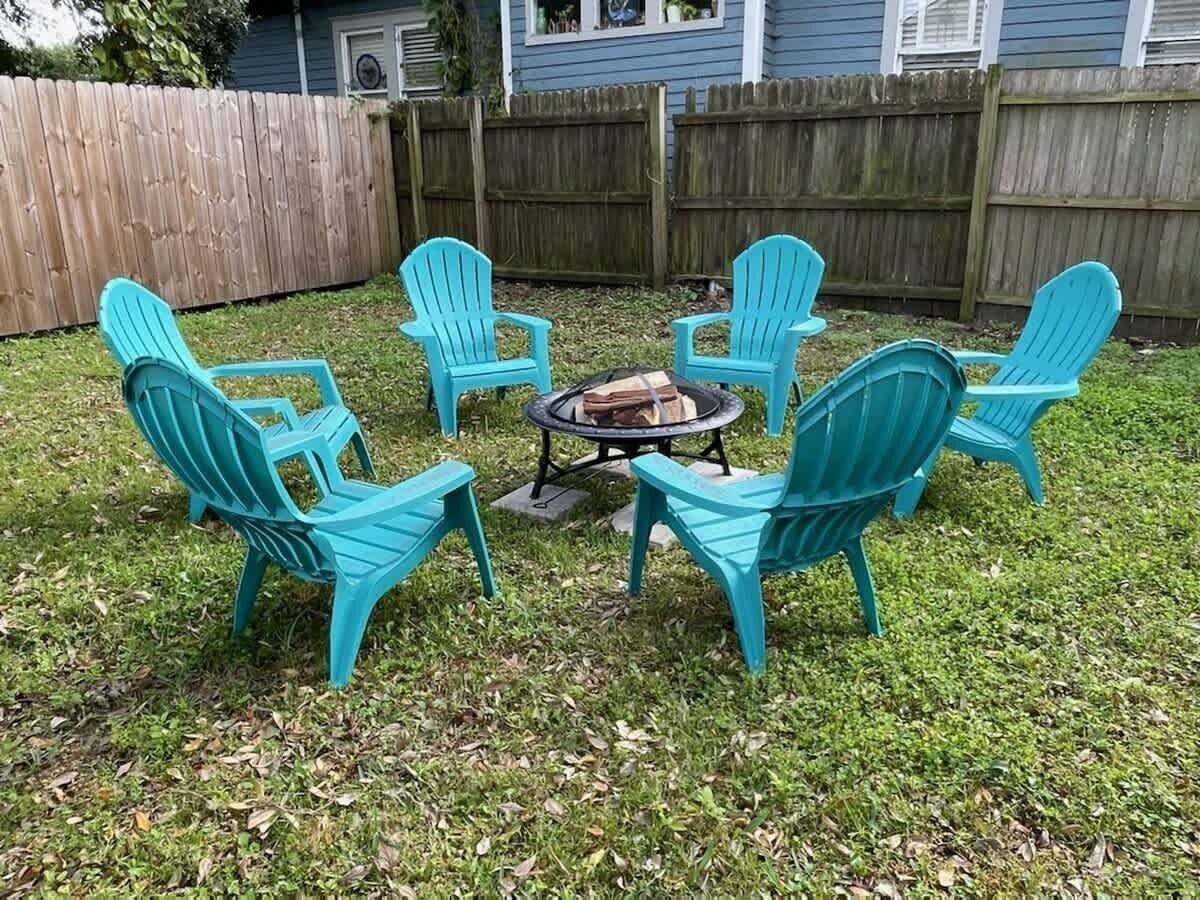 A backyard with a fire pit and six turquoise Adirondack chairs arranged around it.