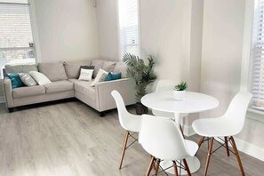 Una sala de estar moderna con un sofá seccional color beige, sillas blancas, una mesita pequeña y una planta en maceta.