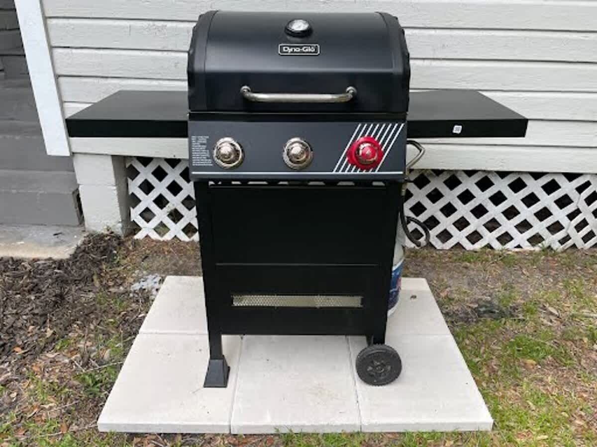 A black outdoor grill with a handle, control knobs, and a red safety lock, placed on a white platform.