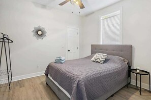 Quarto com cama, ventoinha no teto, janela com persianas, luminária na parede e uma mesinha de cabeceira.