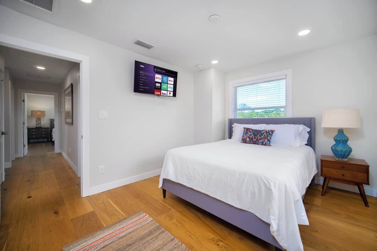 Un dormitorio con cama, televisor montado en la pared, ventana con persianas y mesita de noche con lámpara.