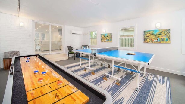 A room with a shuffleboard table, ping pong table, and a pool table. There are paintings on the walls and a chair in the corner. - "Sea Grape Villa" Pool/ Ping Pong, (Jacksonville Beach)