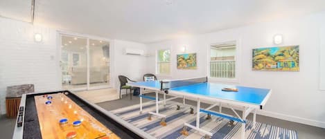 A room with a shuffleboard table, ping pong table, and a pool table. There are paintings on the walls and a chair in the corner.