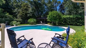 Ein Schwimmbecken mit geschwungener Kante, umgeben von einer Terrasse mit Stühlen und einer kleinen überdachten Struktur.