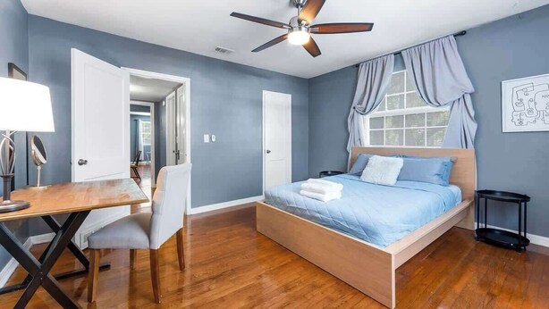 Une chambre à coucher comprenant un lit, un bureau, une chaise, un ventilateur de plafond et une fenêtre avec des rideaux.