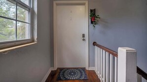 Un couloir avec une porte blanche, une fenêtre, une rambarde en bois et un paillasson.