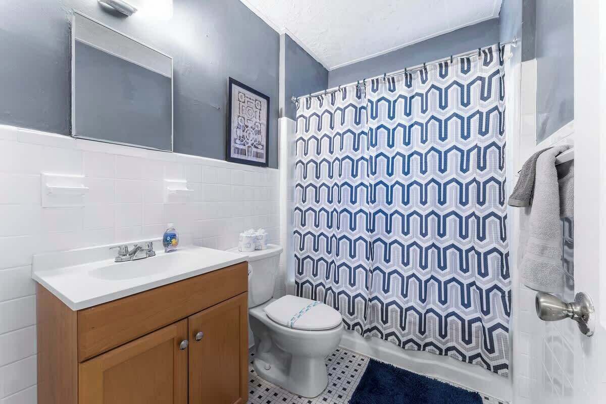 A bathroom with a white sink, wooden cabinet, white toilet, patterned shower curtain, and a framed picture on the wall.