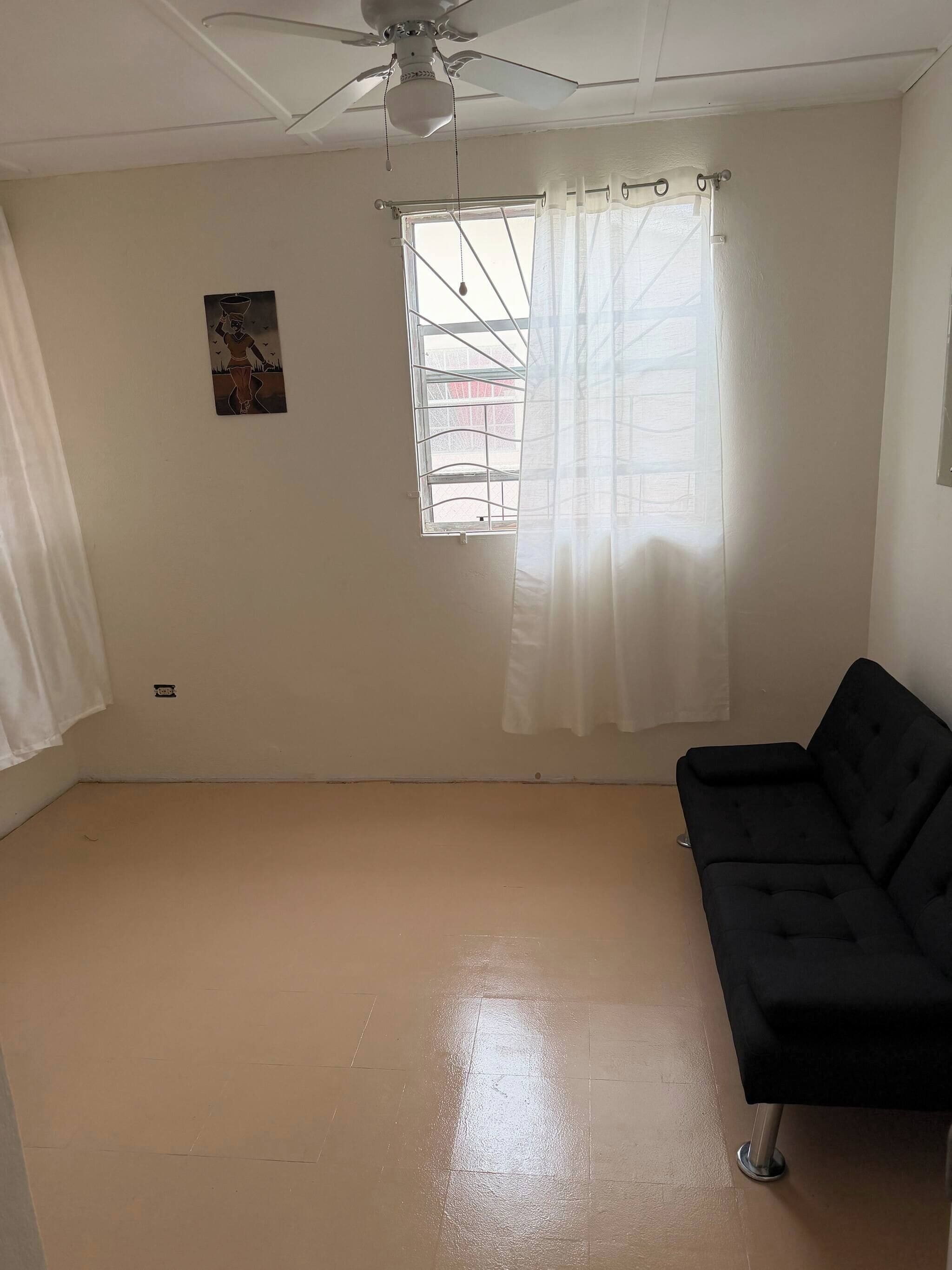 A room with a ceiling fan, a window with a metal grid, a black sofa, and a wall-mounted picture.