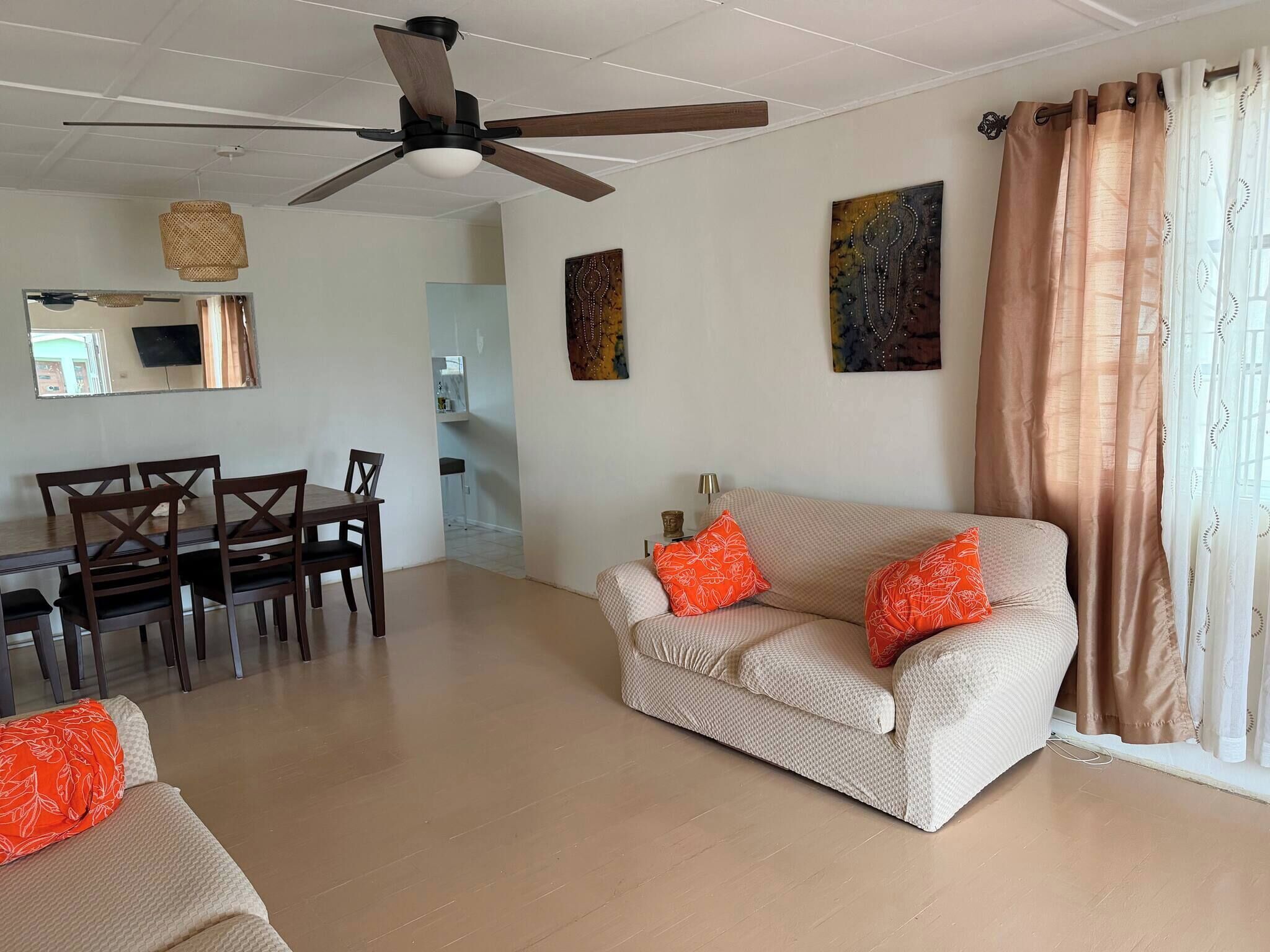 A living room with a ceiling fan, a sofa with orange pillows, a dining table with chairs, and artwork on the walls.