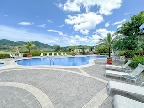 Ein Pool mit Liegestühlen und Topfpflanzen, umgeben von einer Pflasterfläche und Grünflächen.