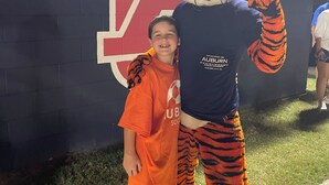 Eine Maskotte in einem Tigerkostüm steht neben einer Person vor einer Wand mit einem großen rot-weißen Logo.