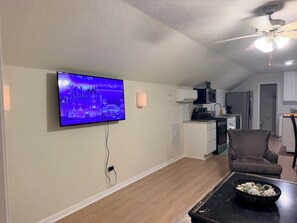 Uma sala de estar com televisão de tela plana na parede, ventilador de teto, área de jantar com mesa e cadeiras, e cozinha com eletrodomésticos.