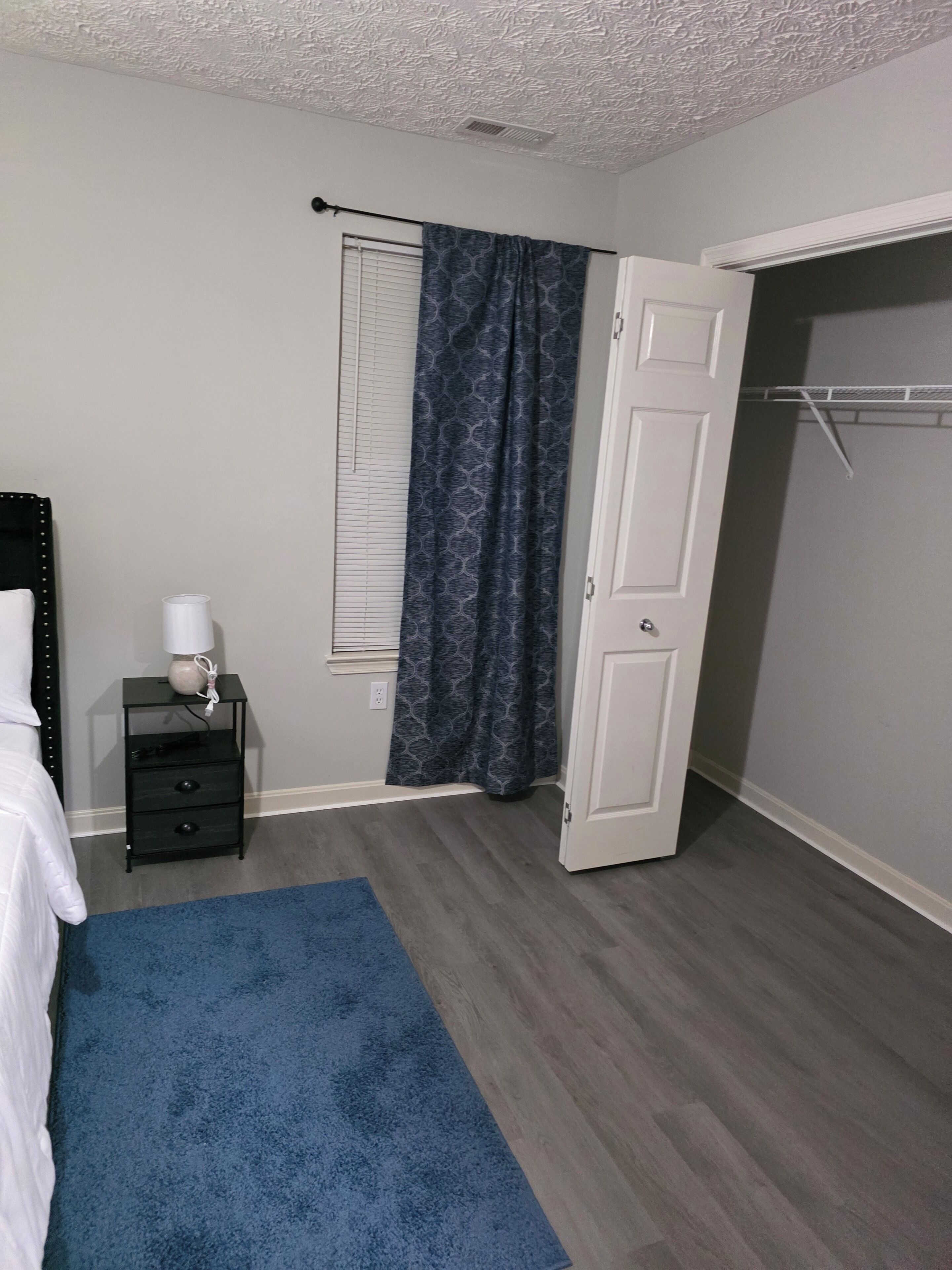 Ein Schlafzimmer mit einem Bett, einem Nachttisch, einem Fenster mit Jalousien, einer Tür und einem blauen Teppich.