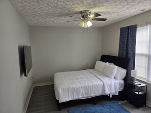 A bedroom with a bed, a nightstand, a ceiling fan, a window with curtains, and a wall-mounted TV.