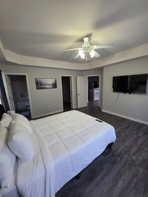 A bedroom with a large bed, a ceiling fan, a flat-screen TV, and a door leading to a bathroom.