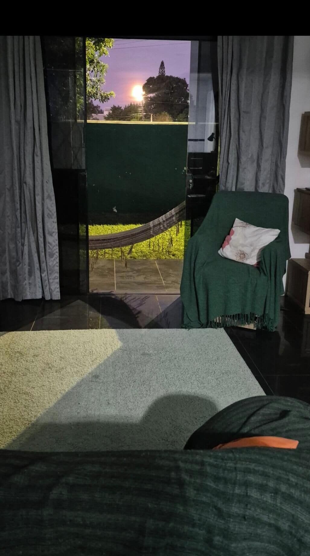 Ein Zimmer mit einem Bett, einem grünen Sessel mit Decke, einer Hängematte und Blick auf einen Sonnenuntergang durch ein Fenster.