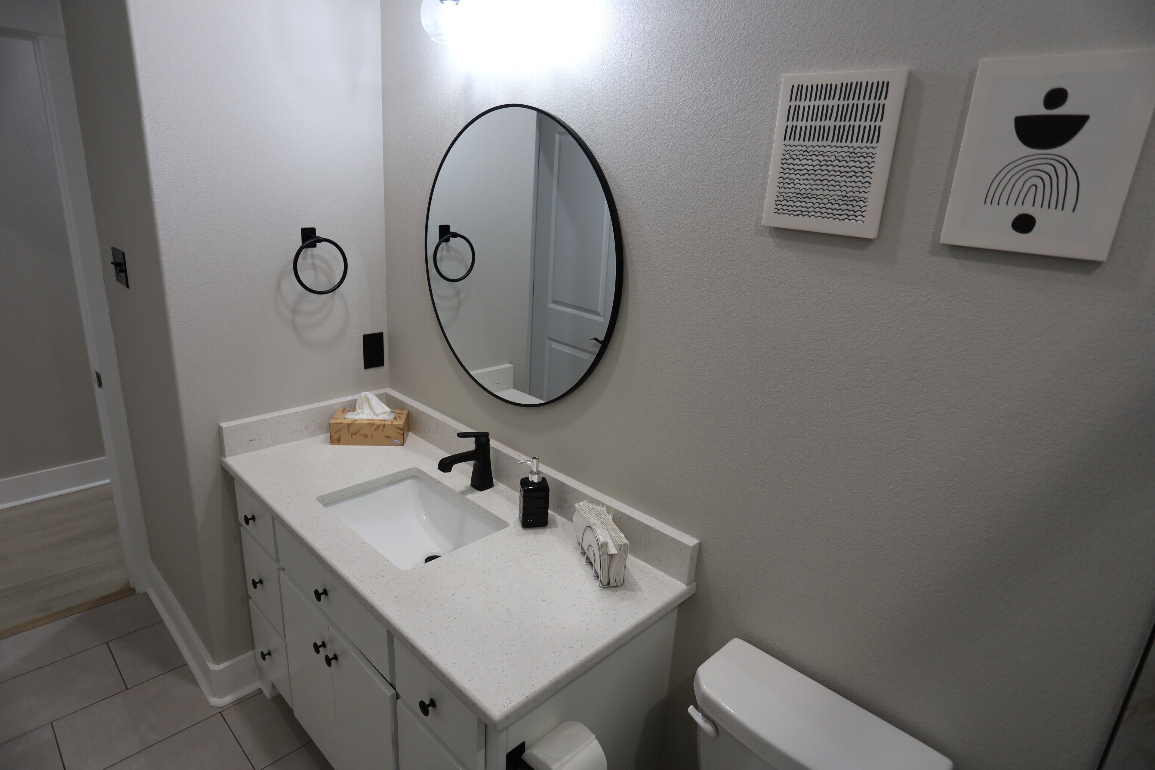 A bathroom with a white sink, black faucet, and a mirror. There are two towel rings on the wall, a toilet, and a white toilet paper holder.