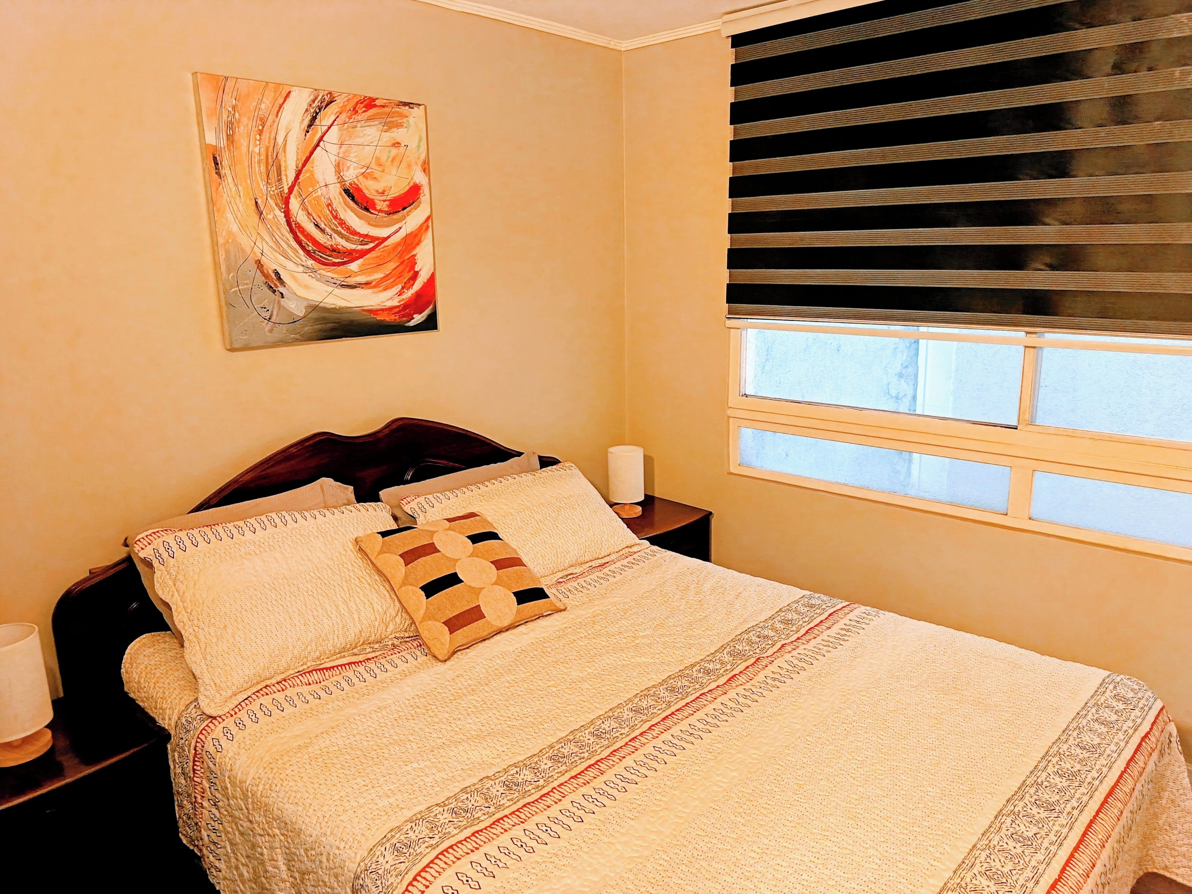 A bedroom with a bed, a nightstand, a lamp, a window with blinds, and a painting on the wall.