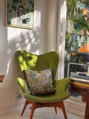 A green armchair with a patterned pillow, positioned next to a window with a view of greenery and a bird feeder.