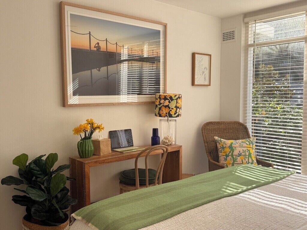 Een slaapkamer met een bed, een bureau, een stoel, een raam met zonwering en een lijst met een afbeelding aan de muur.