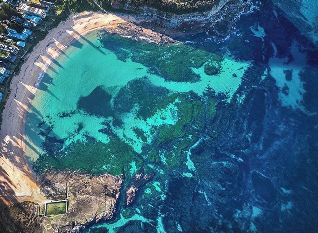 Vue aérienne d’une zone côtière avec une plage, une eau claire et une petite étendue de végétation.