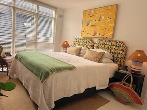 A bedroom with a bed, pillows, a bedside lamp, a nightstand, a woven bag, and a window with blinds.