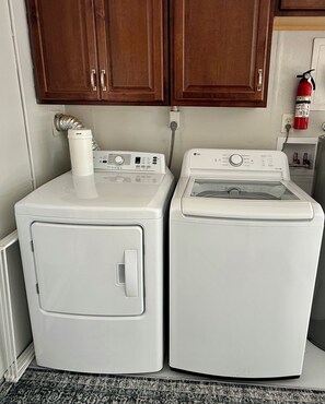A laundry area with two white washing machines and a dryer, a wooden cabinet above, and a fire extinguisher on the wall. - Peace in the City - clean, beautiful 3 BR - private backyard - and near beaches (Sarasota)