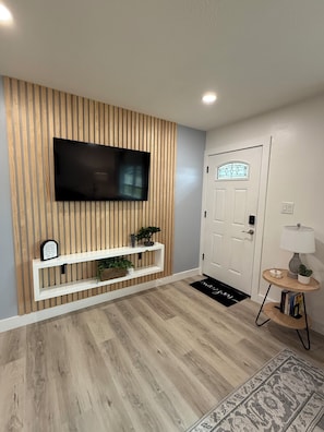 Ein modernes Wohnzimmer mit Wand-Fernseher, Holzpaneelwand, weißem Lowboard, Topfpflanzen, einer weißen Tür und einem kleinen runden Tisch mit Lampe.