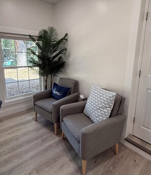 Une pièce avec deux fauteuils gris, une grande plante en pot et une fenêtre donnant sur des arbres.