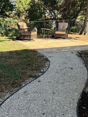 A paved pathway leading to outdoor furniture, surrounded by greenery and a chain-link fence. - Peace in the City - clean, beautiful 3 BR - private backyard - and near beaches (Sarasota)