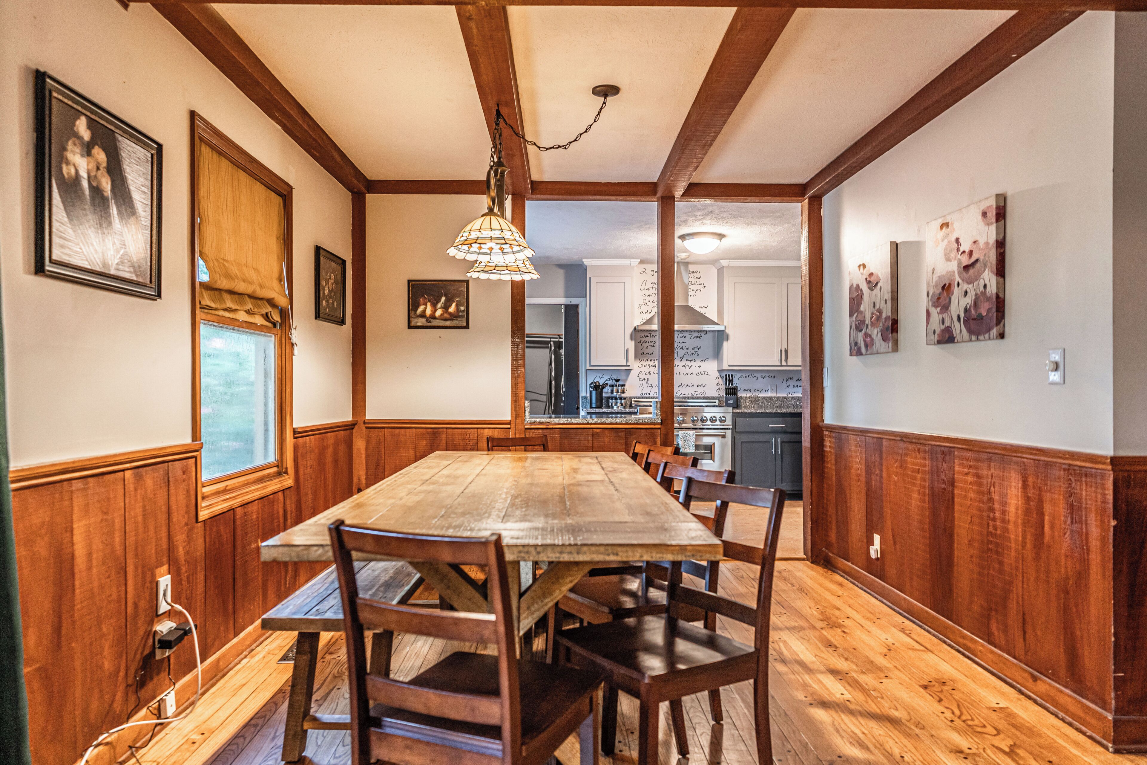 A dining area with a wooden table and chairs, a chandelier, and kitchen in the background.