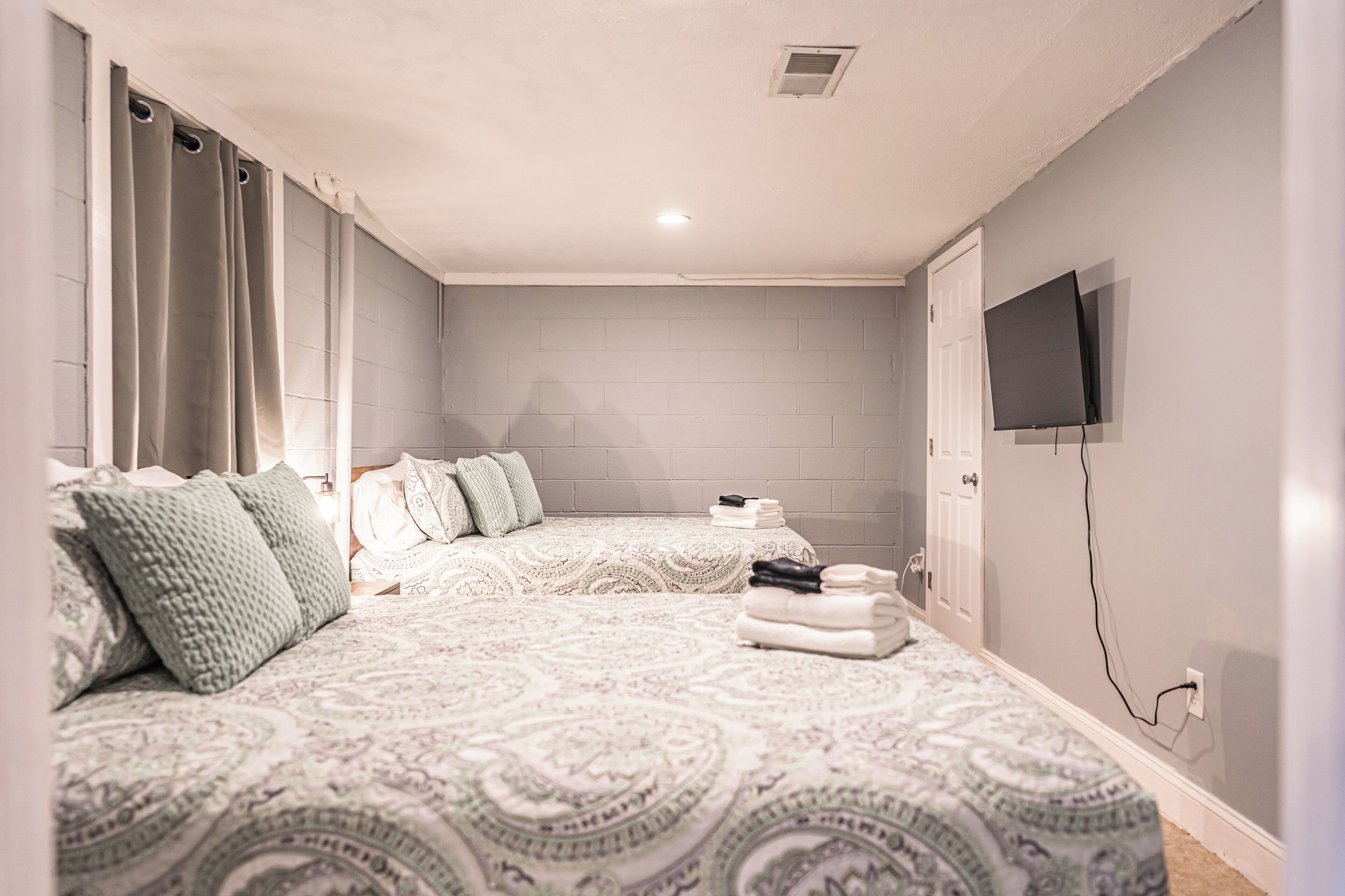 A bedroom with two beds, a flat-screen TV mounted on the wall, and a window with curtains.