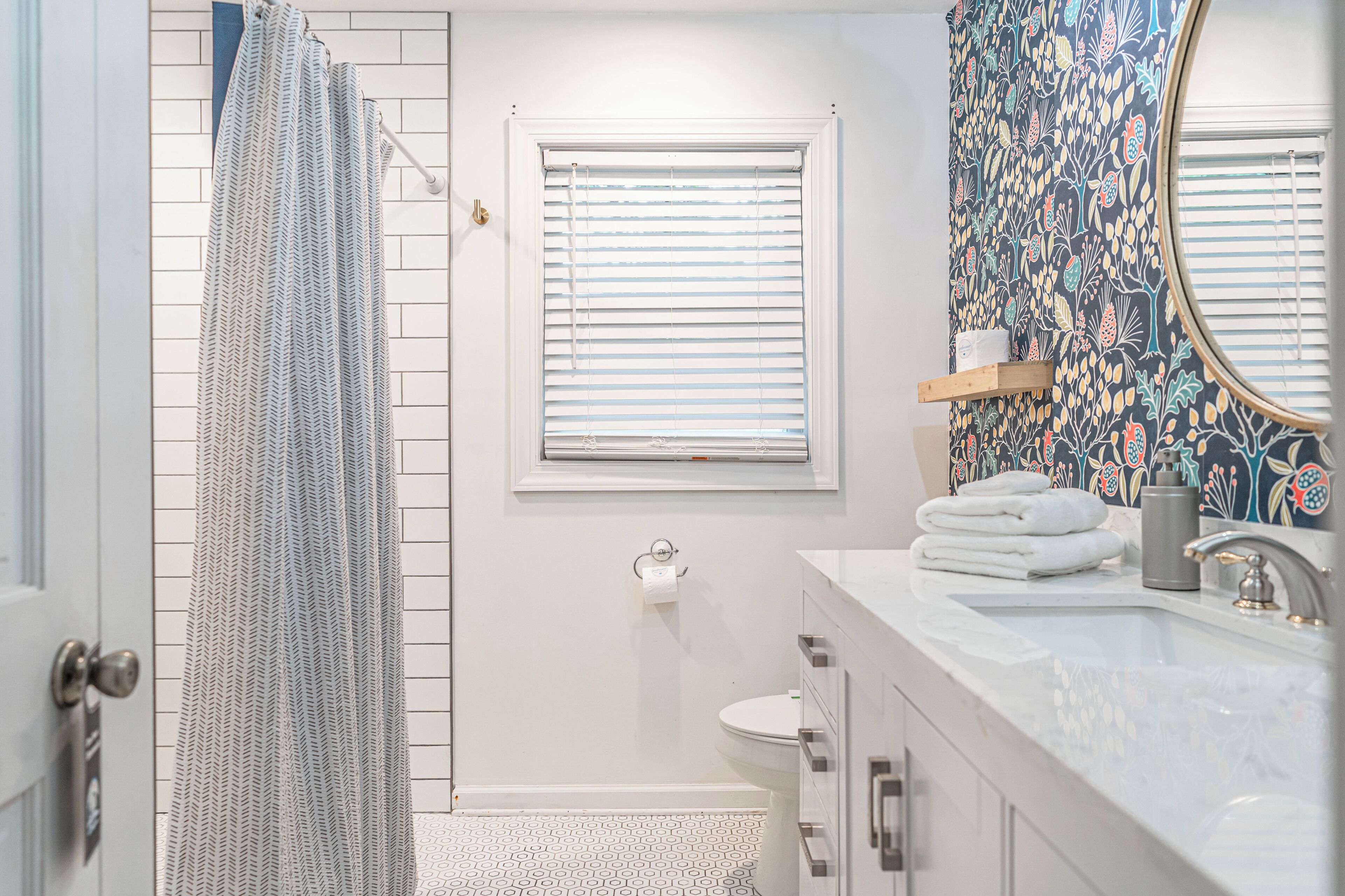 A bathroom with a shower curtain, a toilet, a sink with a mirror, and a window with blinds.