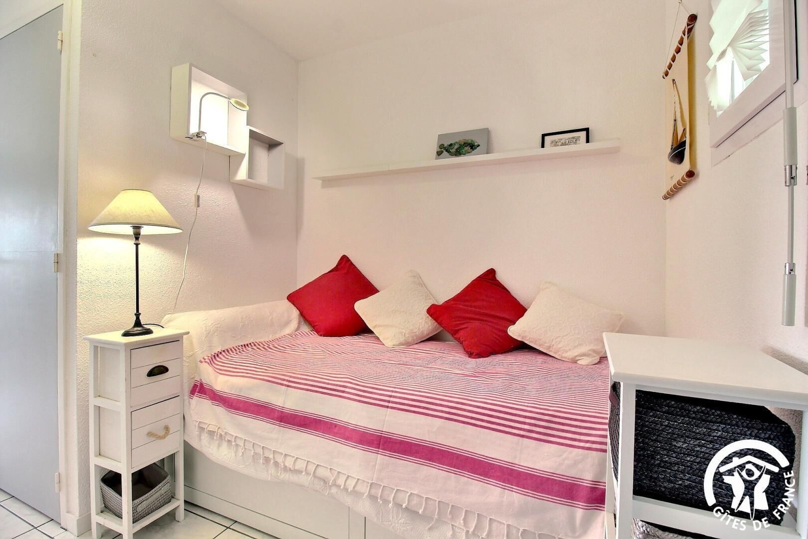 Ein Schlafzimmer mit einem Bett, einem Nachttisch mit Lampe, einer kleinen Schublade, einem Regal mit Dekoration und einem Fenster mit Jalousien.