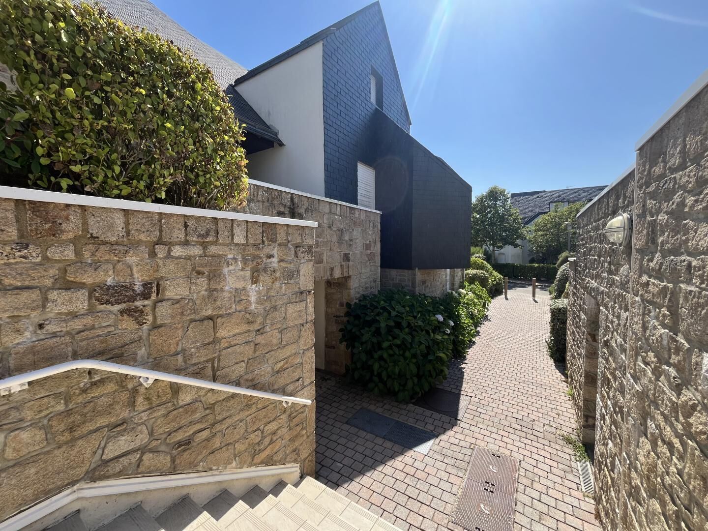 A narrow brick pathway leading to a modern building with a stone wall on one side and a hedge on the other.
