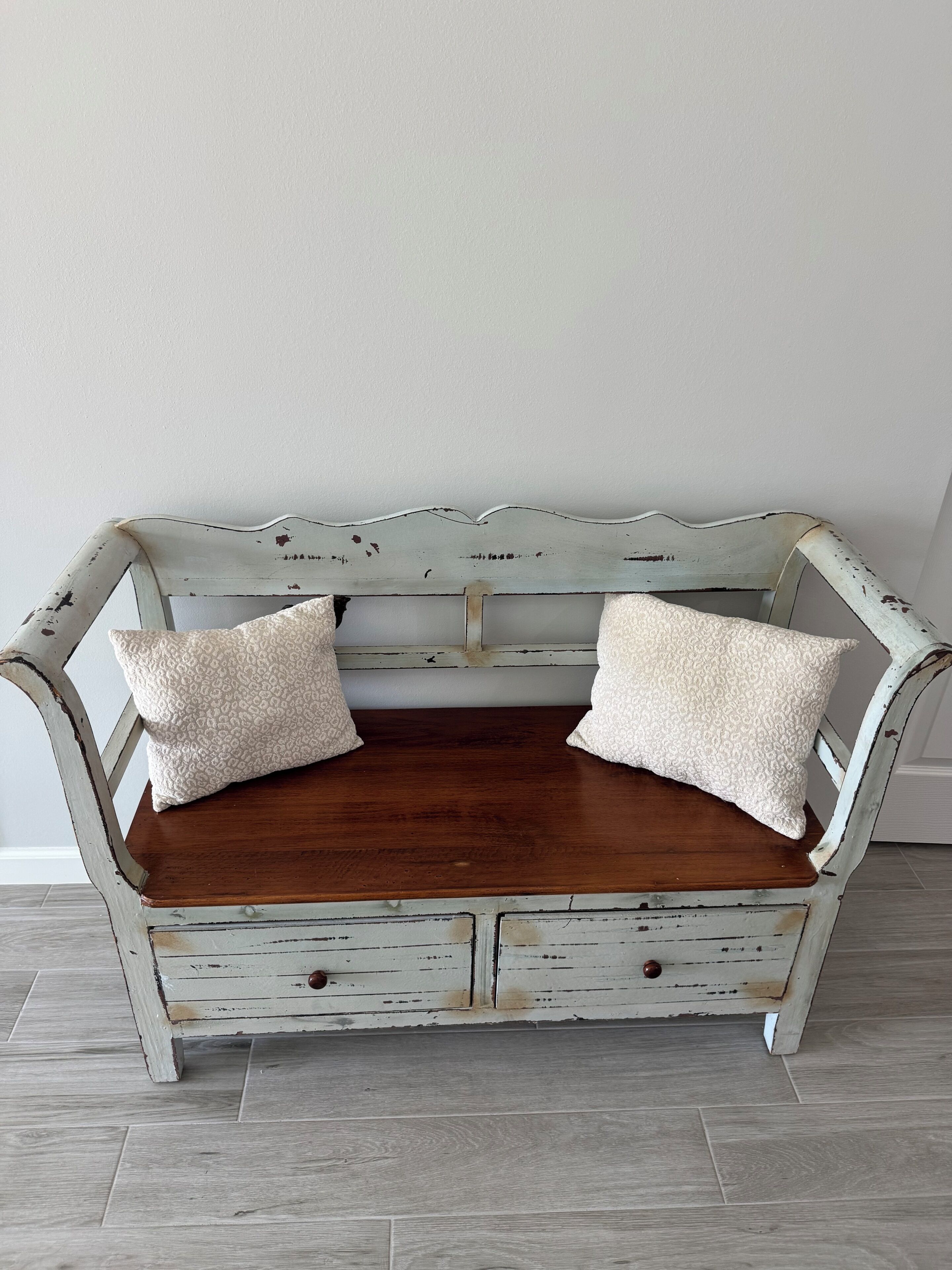 A white, distressed wooden bench with two cushions, a wooden floor, and a plain wall.