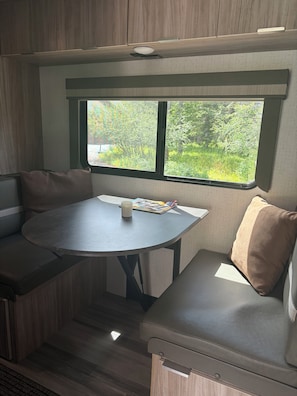 A compact RV interior with a round table, two seating areas, and a window with a view of greenery.