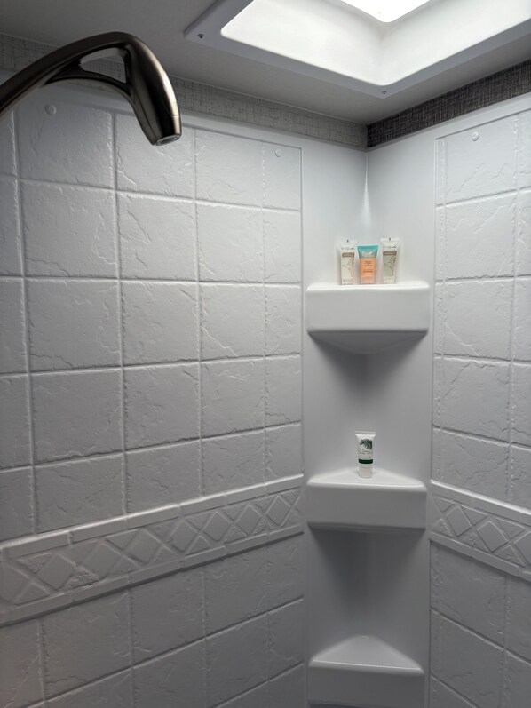 Une salle de bain avec des carreaux blancs à texture, une pomme de douche et une étagère contenant des articles de toilette.