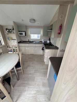 A compact kitchen with a microwave, stove, and sink. There is a dining table with chairs. The room has a light-colored floor and walls.