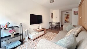 Un salon moderne avec un téléviseur à écran plat, un canapé, une table à manger avec des chaises et un vase décoratif rempli de fleurs.