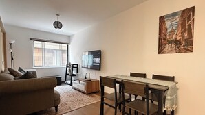 Un salon moderne comprenant un canapé, une table à manger avec des chaises, un téléviseur fixé au mur et une fenêtre avec des stores.