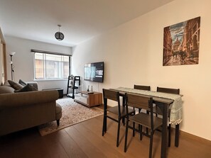 Ein modernes Wohnzimmer mit einer Couch, einem Esstisch mit Stühlen, einem an der Wand montierten Fernseher und einem Fenster mit Jalousien.