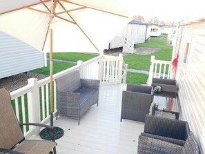 Eine überdachte Terrasse im Freien mit Korbmöbeln, einem weißen Geländer und Blick auf Mobilheime im Hintergrund.