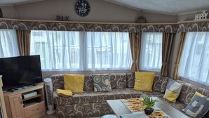 Ein Wohnzimmer mit Sofa, Poufs, einem Tisch mit Pflanze, einem Fernseher und einer Uhr an der Wand.